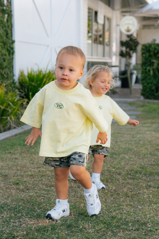 Two children in matching yellow shirts and camouflage shorts running on grass.