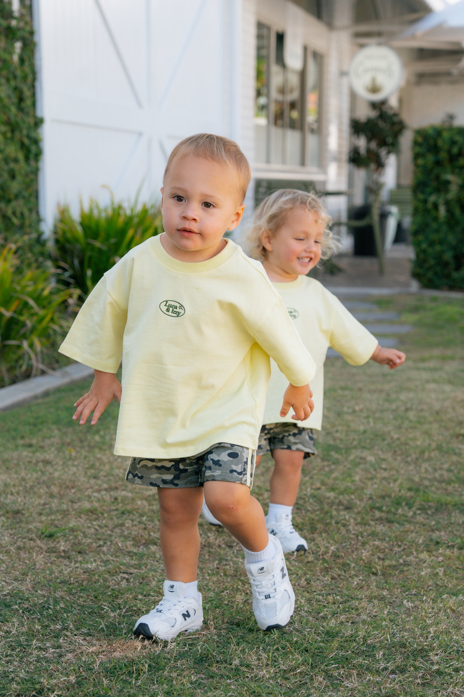 Two children in matching yellow shirts and camouflage shorts running on grass.